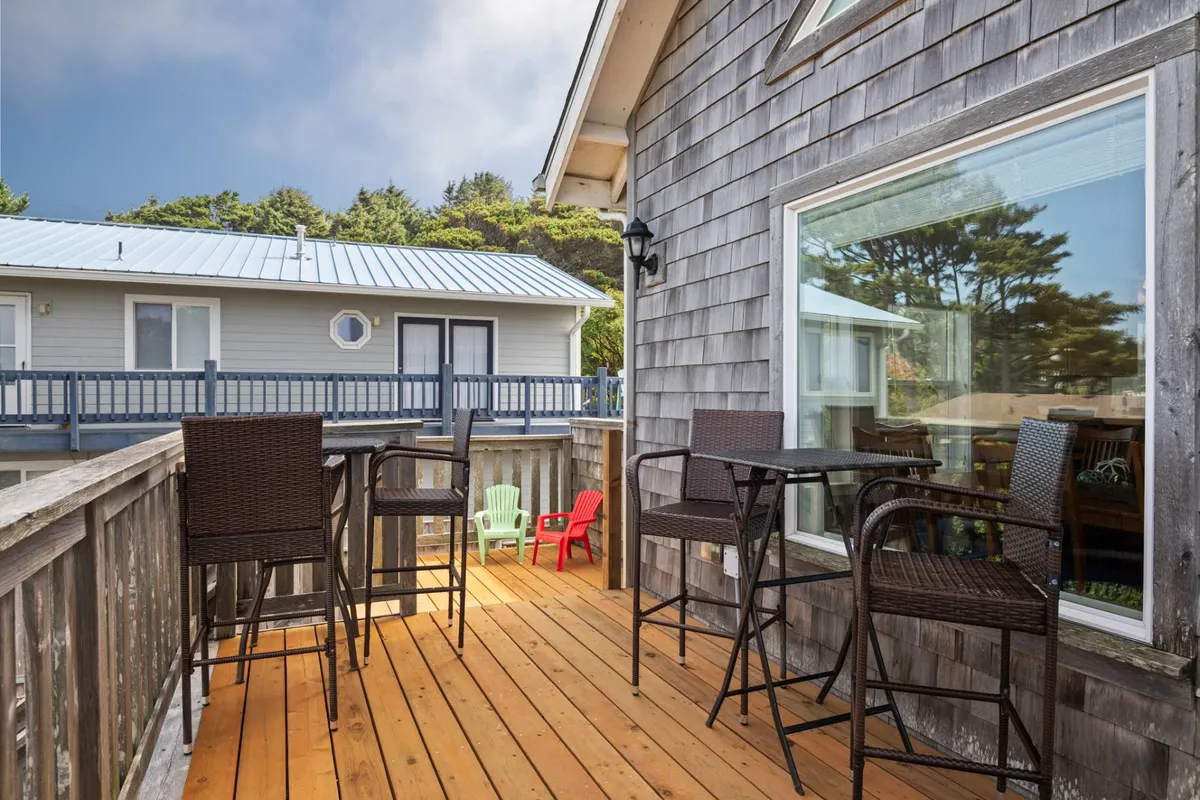 This image shows a wooden deck with high-top outdoor tables and colorful plastic chairs, surrounded by trees and bathed in sunset light. The scene is lively, suggesting a space for socializing, relaxing, and outdoor fun, no matter the weather.