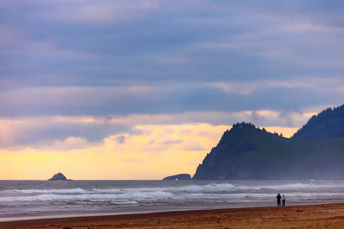 The image shows wide sandy beach with gentle ocean waves, distant cliffs, a glowing sunset sky, and a few people walking along the water’s edge. The scene exudes tranquility and captures the essence of beachside adventure and relaxation.