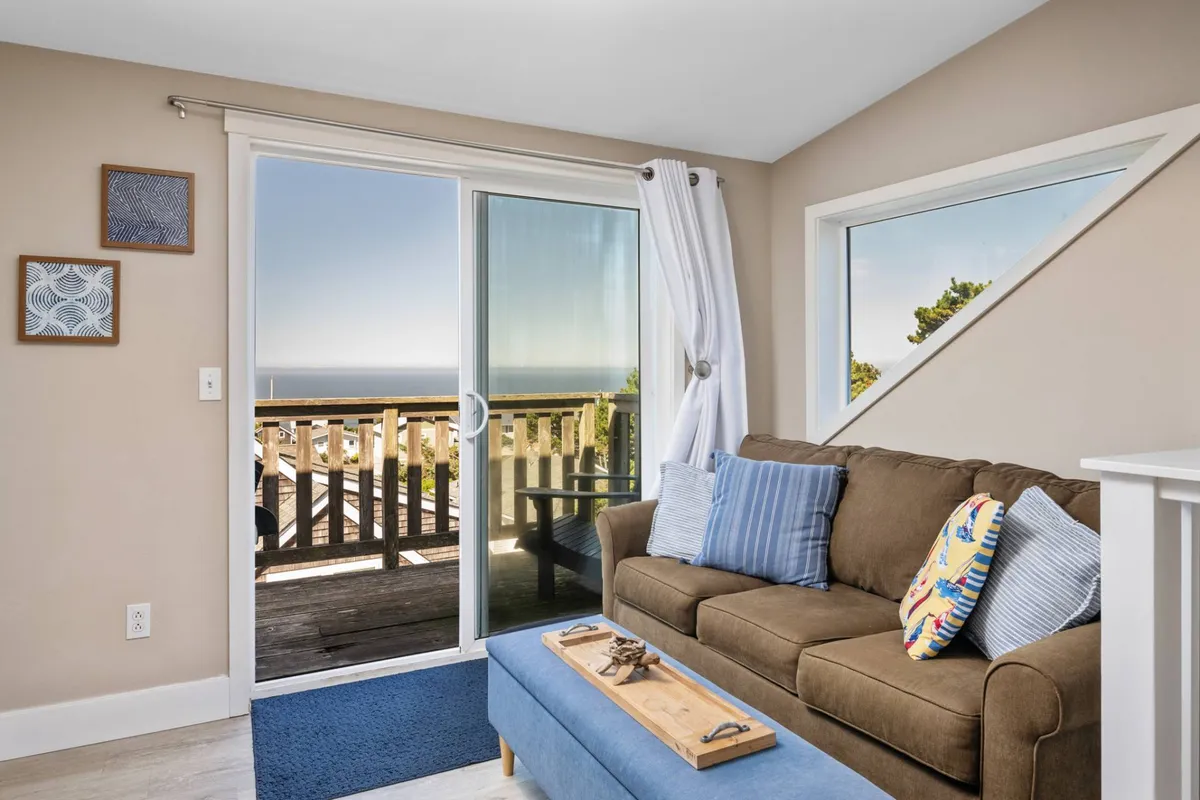 A cozy living room scene with a comfortable couch, coffee table, and large sliding glass door that frames an ocean view. Bright natural light fills the space, and decorative art adds a coastal touch. The sliding door invites thoughts of stepping outside to explore or return from coastal excursions, while the ocean view reinforces the property's proximity to the area's natural beauty.