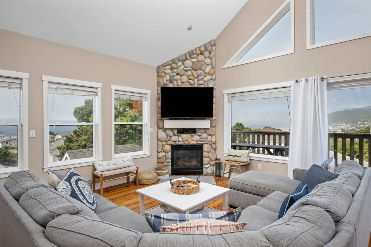 A cozy living room featuring a large stone fireplace, a plush sectional sofa positioned to enjoy both the fire and expansive windows with views, and a coffee table adorned with tasteful decor. The room is filled with natural light, enhancing the warm, welcoming atmosphere.