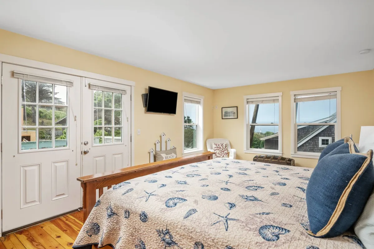 A serene bedroom with a seashell-patterned quilt, large windows framing lush outdoor views, double doors leading directly outside, and a wall-mounted television. The natural light and calming decor create a tranquil, inviting retreat.