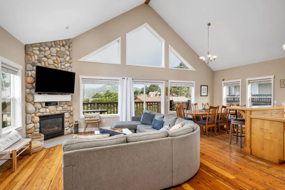 The image showcases a spacious and inviting living area featuring a striking stone fireplace, a plush circular sofa perfect for group gatherings, and expansive windows that capture beautiful sunrise views. The connected dining area is ideal for shared meals and celebrations, with warm light creating a cozy, welcoming atmosphere.