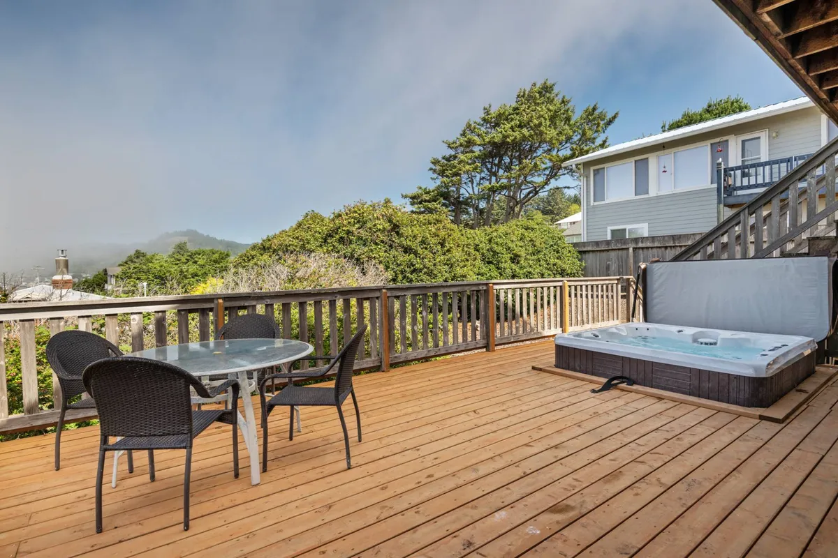 A wooden deck surrounded by lush greenery, featuring a hot tub and a spacious outdoor dining table. The area is inviting and tranquil, ideal for gatherings, alfresco meals, and peaceful moments in nature.