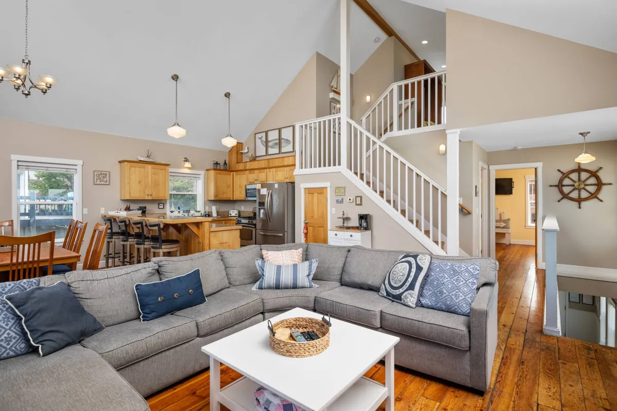 A spacious, open-concept main level combining a comfortable sectional sofa, a substantial wooden dining table, and a modern kitchen area. The layout flows seamlessly, with a staircase visible, highlighting the communal nature of the space.