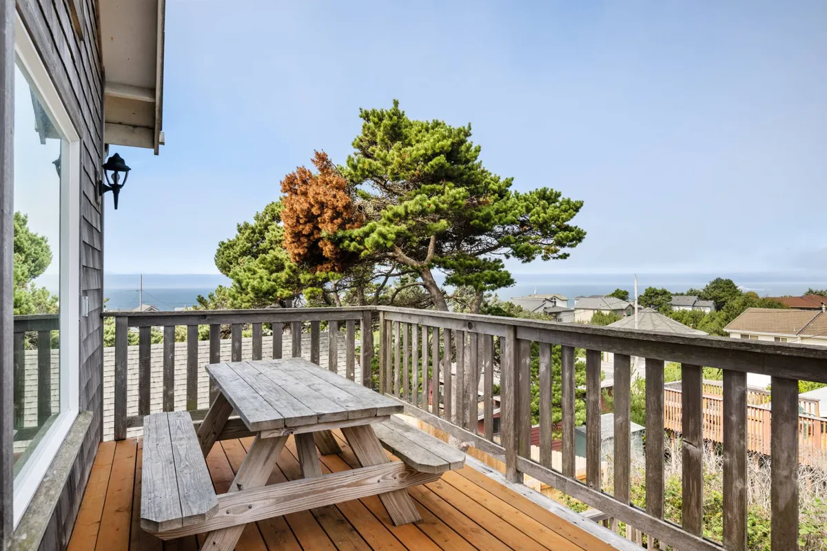 A wooden picnic table sits beneath lush green trees with a clear view of the ocean beyond. The scene is tranquil, inviting guests to relax, dine outdoors, or simply enjoy the sights and sounds of the coastline.