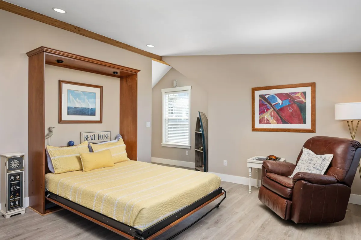 A bright loft space with a foldable Murphy bed, a comfortable recliner chair, vibrant artwork, and a large window letting in natural light. The flexible setup invites both daytime relaxation and restful sleep at night.