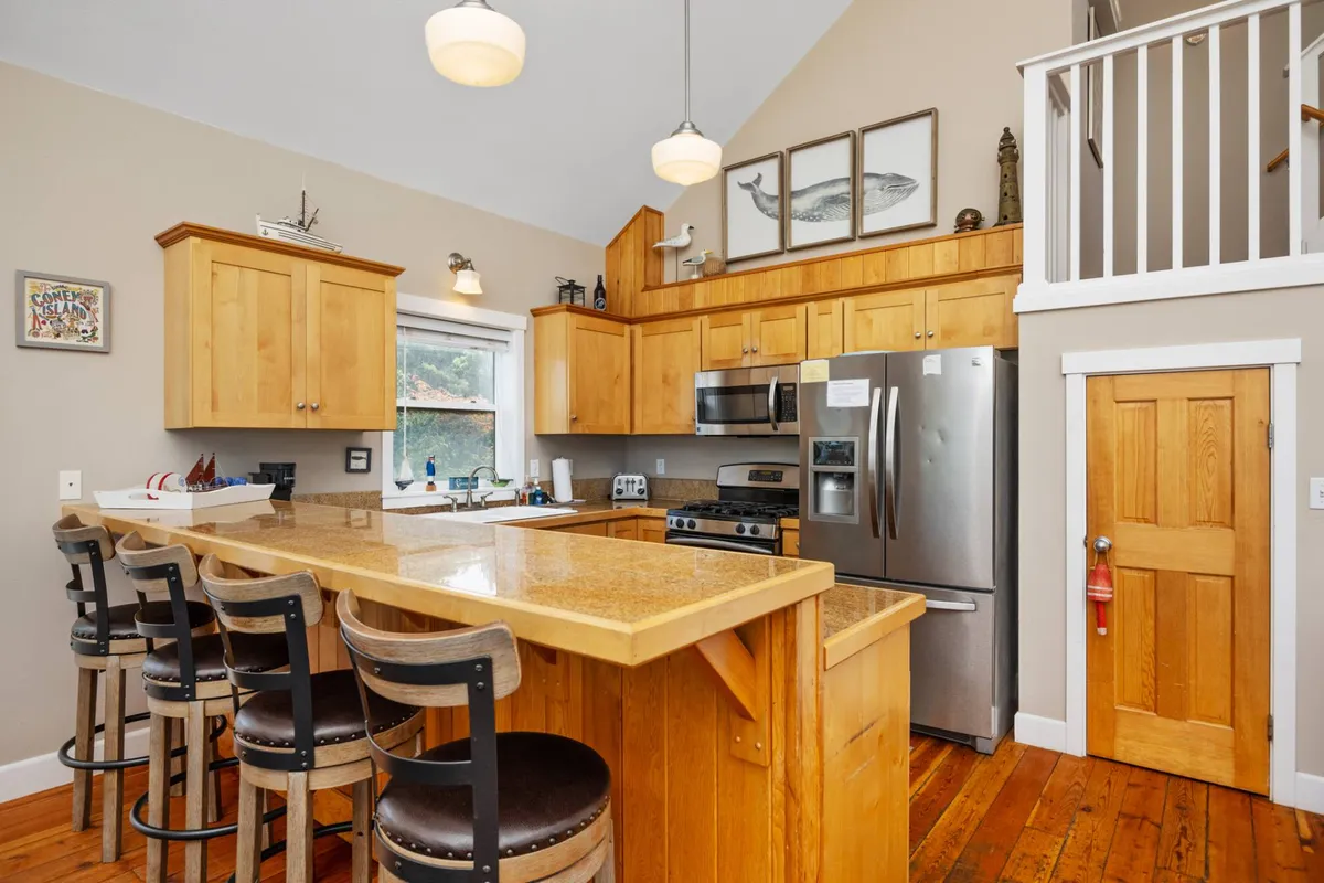 A stylish, modern kitchen with granite countertops, wooden cabinetry, bar stools at a breakfast bar, and stainless steel appliances. The setting is ready for group cooking and casual dining, with inviting decor and ample space.