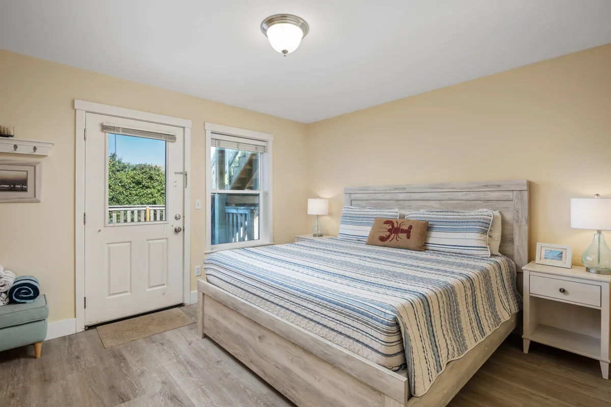 A bedroom featuring striped bedding, a door leading directly outside, a window opening to lush greenery, and a cozy nightstand with a lamp. The layout emphasizes easy access to the outdoors and the comfort of returning to a serene retreat after an active day. The natural light and outdoor connection invite guests to step out and start their adventures early each morning.